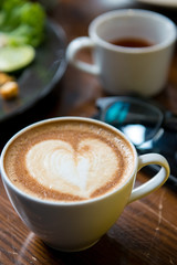 A cup of Cappuccino coffee put on wooden table with glasses and tea in background.