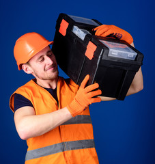 Worker, repairer, repairman, strong builder on cheerful smiling face carries toolbox on shoulder, ready to work. Repair service concept. Man in helmet, hard hat holds toolbox, blue background.