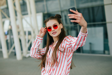 Pretty fashion model in red glasses takes selfie on her smartphone standing before a modern glass building