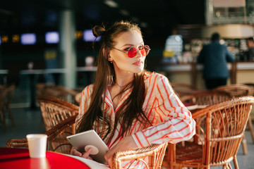 Pretty fashion model in red sunglasses works with her tablet sitting at the cafe in the rays of sunset