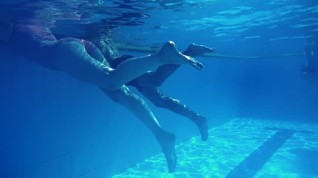 Woman and man in swimsuits simultaneously raise their feet in pool. Swim in the water. Sexually move your legs under water. Slender body. Underwater shooting. Slow motion.