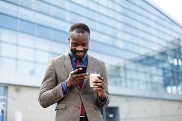 Happy African American man looks lucky reading something in his smartphone while he stands outside with a cup of coffee