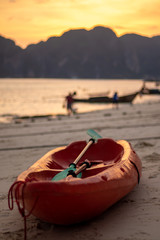 A red canoe on a beach with a golden hour sunset over the mountains in the background. The beach is Long beach on Koh Phi Phi island along the Andaman coast of Thailand.