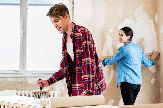 Young man applying adhesive to the back of the wallpaper sheet while his girlfriend is measuring the prepared wall in their new home