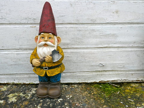 Gartenzwerg mit rotem Hut, gelber Jacke, blauer  Hose und braunen Stiefeln. Er hat eine Axt in der Hand und steht vor einer verwitterten, weißen Bretterwand