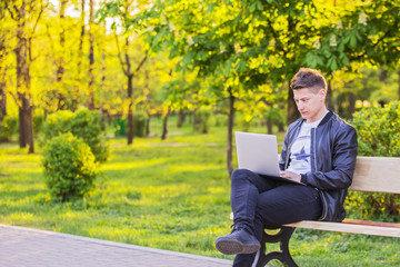 A young handsome man is sitting and working in the park with a laptop. The guy freelancer works outside