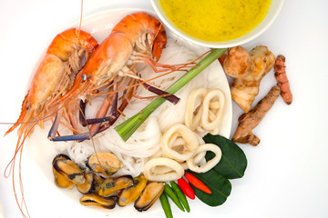 Top view : Rice vermicelli and shrimps, squid and mussel are in white plate. Seafood curry in white bowl. Chillies, ginger, turmeric and lime leaf on white background.