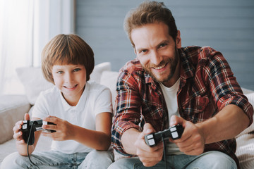 Cheerful father with game controller plays video game with small son sitting on couch. © VadimGuzhva