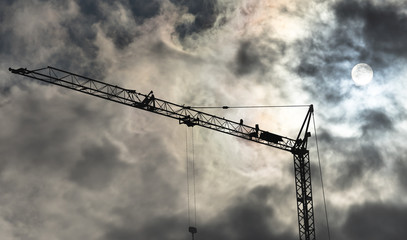 High black construction crane in front of a dramatic threatening sky with translucent sun behind the clouds