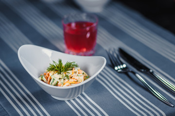Table in the restaurant. On the table is a salad in a white plate, a knife, a fork, a glass with red juice. On the table is a blue tablecloth in strips.