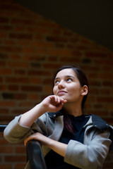 Indoor portrait of young beautiful model of mixed race dressed in gray coat standing at the staircase of brick building