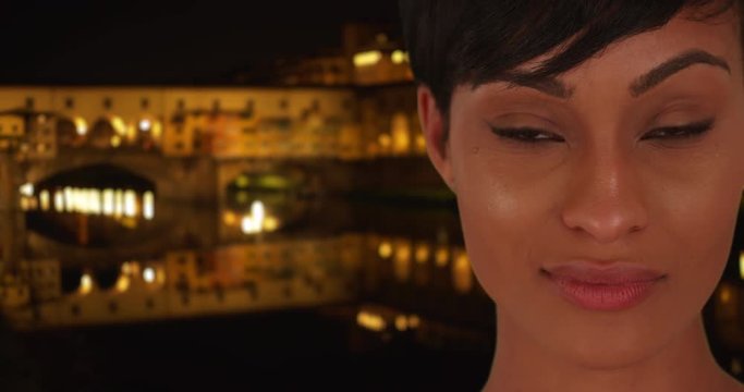 Beautiful young black female stares deeply at the camera while standing outdoors in Florence near the Ponte Vecchio, Gorgeous African woman looking longingly at the viewer at night in Italy, 4k
