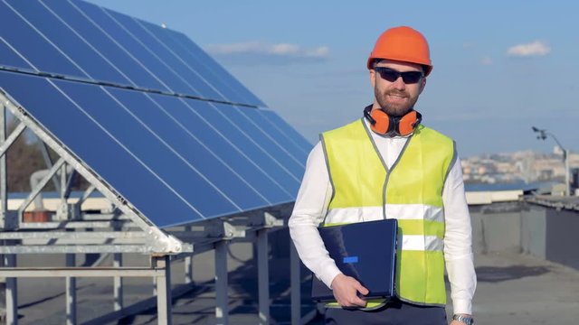 A worker smiles at camera, while standing near solar panels and holding a laptop. 4K.