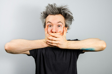 Unshorn and unshaven young guy with piercings on his face, closing his mouth with his hands on gray...