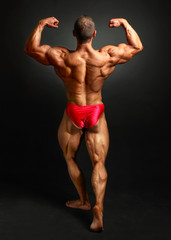 Fototapeta premium Young bodybuilder man flexing back double bicpes pose, showing his huge trapezius, rhomboid, levator, latissimus dorsi and legs muscles. Studio shot on black background.