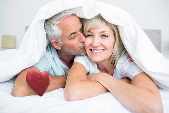 Closeup of mature man kissing womans cheek in bed against red heart