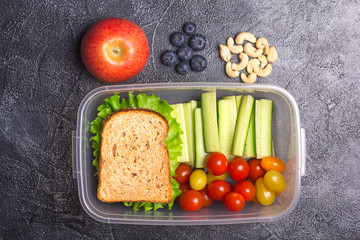 Healthy lunch box with sandwich and fresh vegetables and red apple, blueberries, cashew on dark stone table. Top view