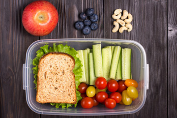 Healthy lunch box with sandwich and fresh vegetables and red apple, blueberries, cashew on brown rustic wooden table. Top view