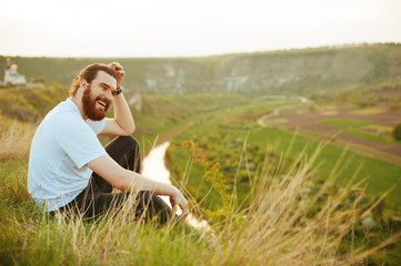 Naklejka na ściany i meble Happy bearded man sitting on grass in nature outdoor and smiling at sunset