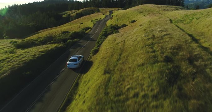 Aerial view of car driving down country road through rural rolling hills at sunset