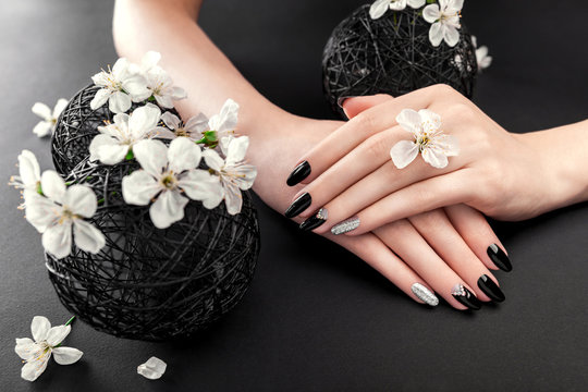 Black and silver manicure with cherry blossom on black background. Woman with black nails surrounded with white flowers