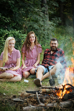 Smiling friends sitting next to campfire. Happy youngsters camping in forest. Bearded guy lying on grass while girls read books