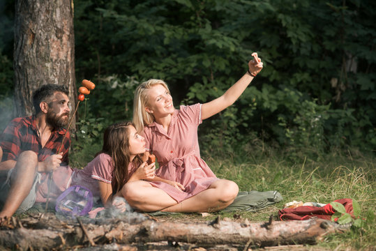 Summer picnic in woods. Smiling blond girl taking selfie in forest or park. Cheerful friends posing with sausages for photo