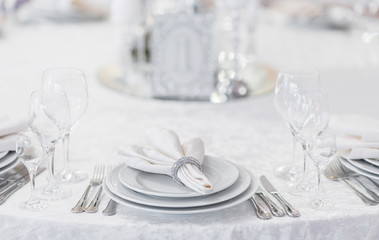 Silver cutlery and stylish white plates are laid out on a white tablecloth of expensive fabric in a restaurant for a festive banquet - obrazy, fototapety, plakaty