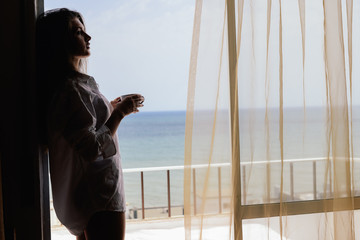 Cheerful beautiful woman enjoying healthy relax lifestyle drinking hot beverage looking out window having positive day and bright sunny oceanfront outdoors background. Travel relaxation destination