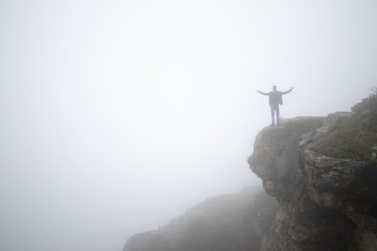 Hombre joven moreno con barba de pie con los brazos abiertos en el borde de la montaña un día de niebla y mal tiempo