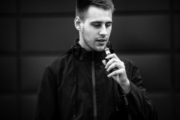 Fototapeta na wymiar Vape teenager. Handsome young white guy with modern haircut in checkered sleeveless shirt vaping an electronic cigarette opposite the futuristic urban background in the spring. Black and white.