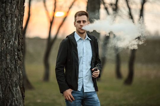 Vape teenager. Handsome young white guy in black jacket and checkered shirt vaping an electronic cigarette among the trees in the park at sunset. Lifestyle.