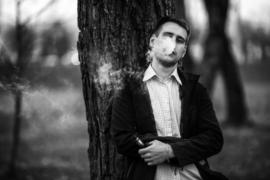 Vape teenager. Handsome young white guy in black jacket and checkered shirt vaping an electronic cigarette among the trees in the