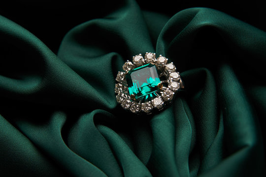 Green emerald fashion engagement diamond ring on green satin background. Luxury female jewellery, close-up. Selective focus