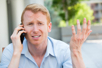 Closeup portrait, worried young man in blue shirt talking on phone to someone, looking gloomy, isolated outdoors outside background