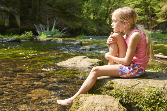  Child blond girl sitting on a rock by the river summer sunlight