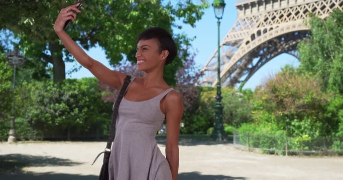 Cheerful young mixed race female taking selfies on her smartphone near the Eiffel Tower in Paris, Playful millennial traveling in France female taking photos of herself, 4k