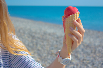 Female hand holding a red ice cream in a waffle cone on background of blue sea. Summer vacation concept