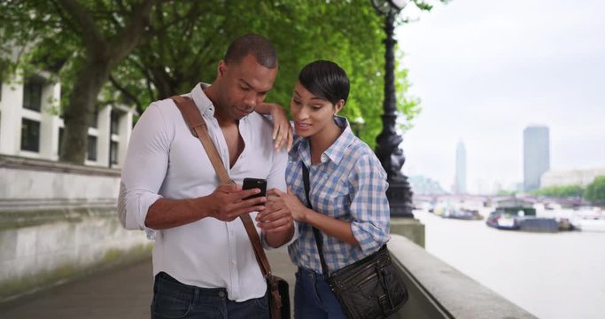 Young loving ethnic couple watch something amusing together on a smartphone while walking around in London, African-American man and woman using a smartphone outside, 4k