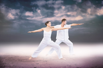 Peaceful couple in white doing yoga together in warrior position against dark cloudy sky