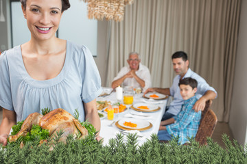 Woman holding chicken roast with family at dining table against green fir branches