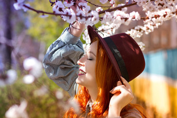 Stylish young beautiful smiling red-haired girl in a burgundy hat near a flowering tree - 203176112