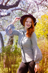 Stylish young beautiful smiling red-haired girl in a burgundy hat near a flowering tree - 203176133