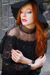 Stylish young beautiful red-haired girl in a black hat near the stone wall