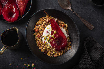 homemade granola (hazelnuts, raisin, dried cranberry, pumpkin, sesame and sunflower seeds) with yogurt and poached pear in black bowl.