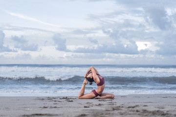 side view of young woman practicing yoga in One Legged King Pigeon pose (Eka Pada Rajakapotasana) on seashore