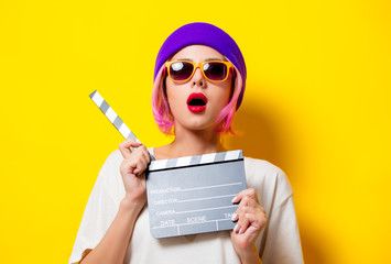 Young girl with pink hair in purple hat and sunglasses holding movie clapper on yellow background