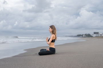 Fototapeta na wymiar side view of beautiful young woman practicing yoga in lotus pose (padmasana) with namaste mudra on seashore