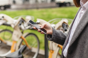 Close-up shot of hands using smartphone, young girl using bicycle rent mobile app smiling outdoors, female manager browsing smartphone standing near bike sharing, healthy city lifestyle