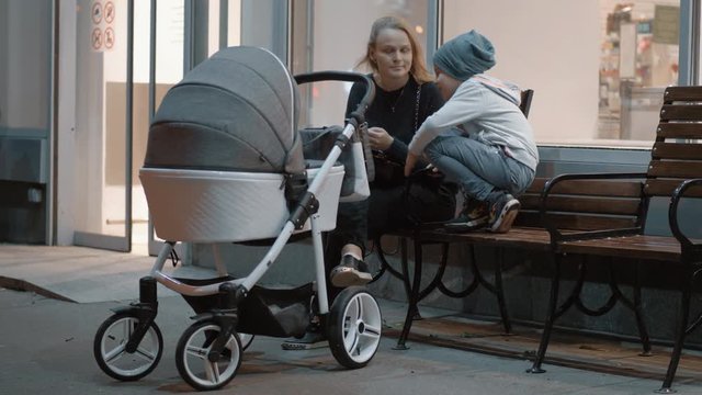 Family spending evening together outside. Mum talking to son sitting on the bench near the store while baby is sleeping in pram